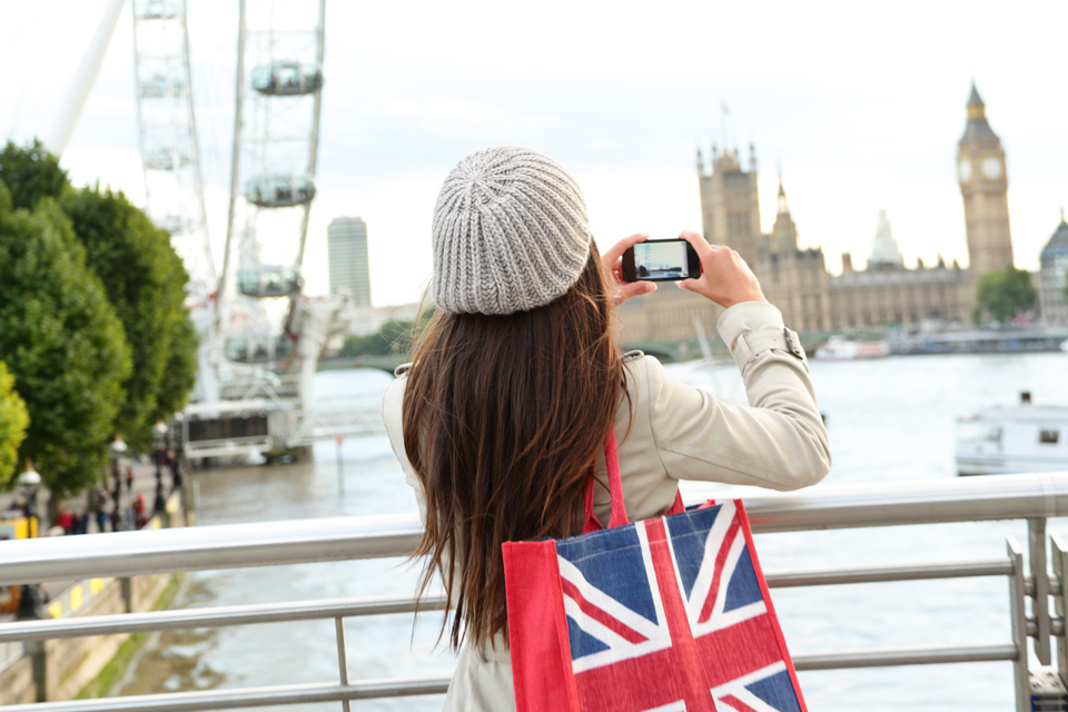 A women, with long dark hair, holding a Union Jack bag, taking a photo of the Houses of Parliament and Big Ben. 